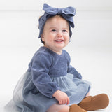 A smiling baby with light hair sits on the floor in a Mabel and Honey Skylar Blue Suede Dress, styled with a tulle skirt, corduroy bodice, a matching blue bow headband, and brown shoes against a plain white background.