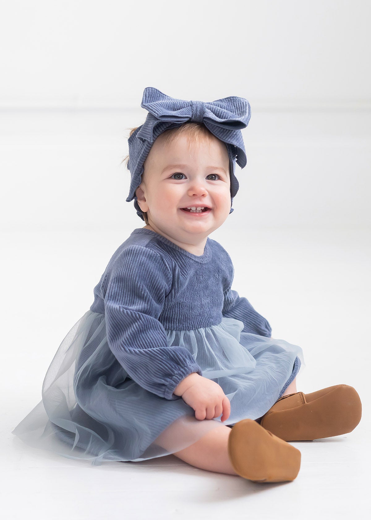A smiling baby with light hair sits on the floor in a Mabel and Honey Skylar Blue Suede Dress, styled with a tulle skirt, corduroy bodice, a matching blue bow headband, and brown shoes against a plain white background.