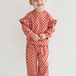 A young child with light brown hair in two buns smiles while wearing the Mabel and Honey Red Velvet Two Piece Set, featuring pink knit fabric with red polka dots and ruffled sleeves, plus white sneakers, against a plain light background.