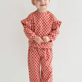 A young child with light brown hair in two buns smiles while wearing the Mabel and Honey Red Velvet Two Piece Set, featuring pink knit fabric with red polka dots and ruffled sleeves, plus white sneakers, against a plain light background.