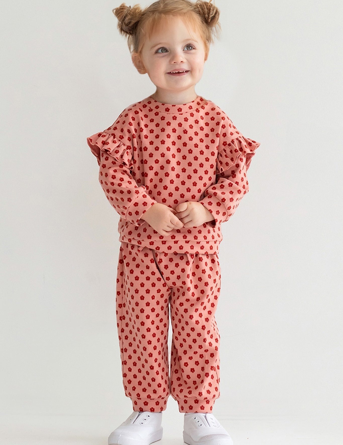 A young child with light brown hair in two buns smiles while wearing the Mabel and Honey Red Velvet Two Piece Set, featuring pink knit fabric with red polka dots and ruffled sleeves, plus white sneakers, against a plain light background.