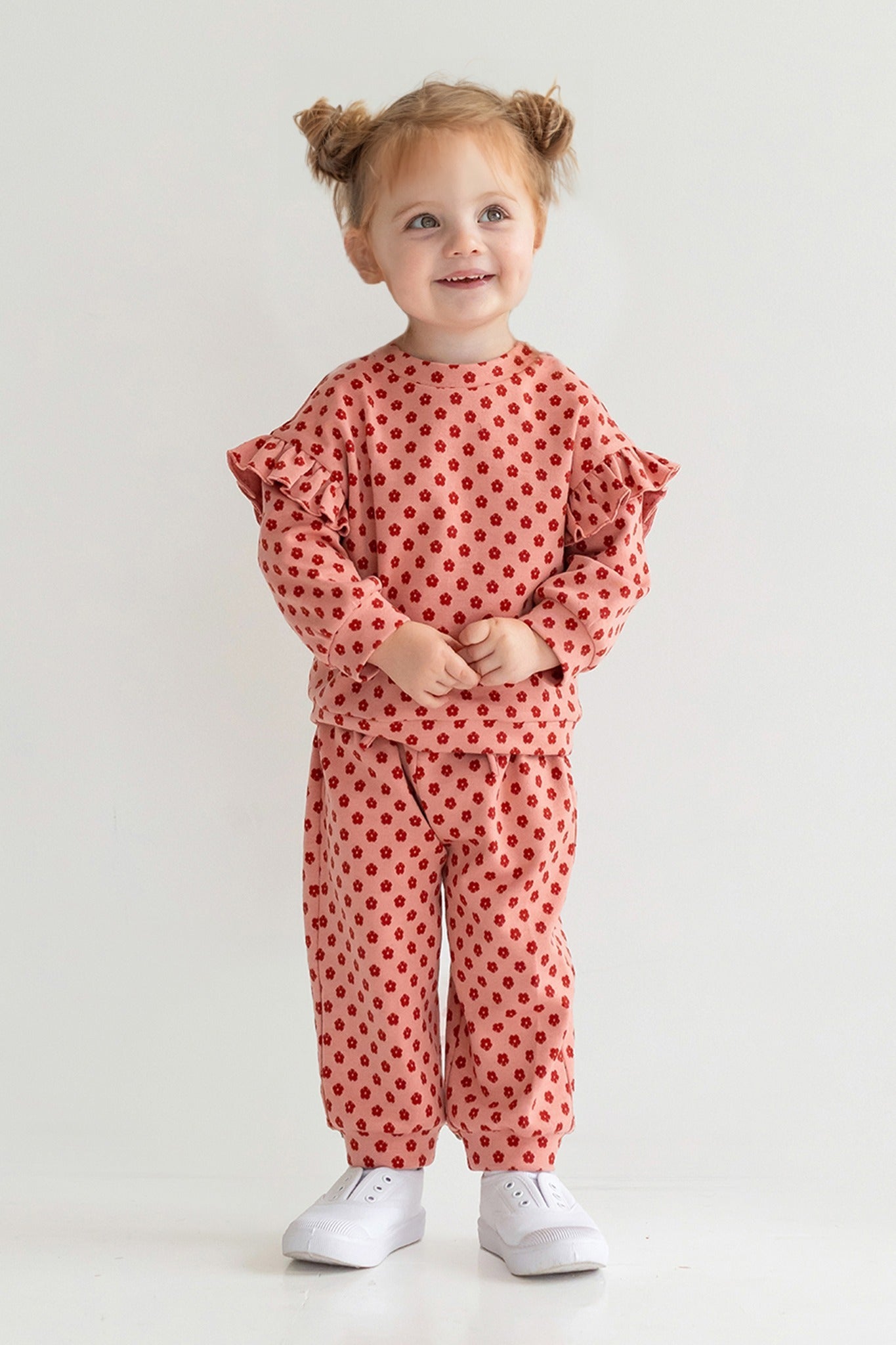 A young child with light brown hair in two buns smiles while wearing the Mabel and Honey Red Velvet Two Piece Set, featuring pink knit fabric with red polka dots and ruffled sleeves, plus white sneakers, against a plain light background.