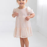 A young girl with light brown hair in two buns smiles in a bright, minimalist room, wearing the Taylor Sequined Toddler Dress by Isobella and Chloe—light pink with puffed sleeves—paired with silver shoes.