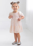 A young girl with light brown hair in two buns smiles in a bright, minimalist room, wearing the Taylor Sequined Toddler Dress by Isobella and Chloe—light pink with puffed sleeves—paired with silver shoes.