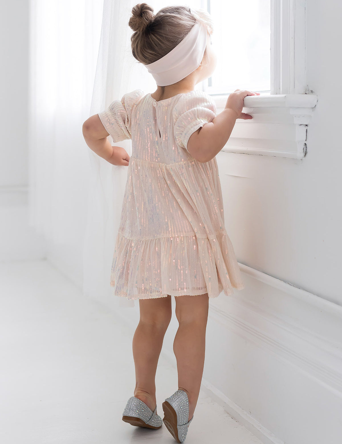A young girl wears the Isobella and Chloe Taylor Sequined Toddler Dress, paired with silver shoes and a white headband, standing on tiptoes by a bright window in a softly lit white room, her hair styled in a bun.