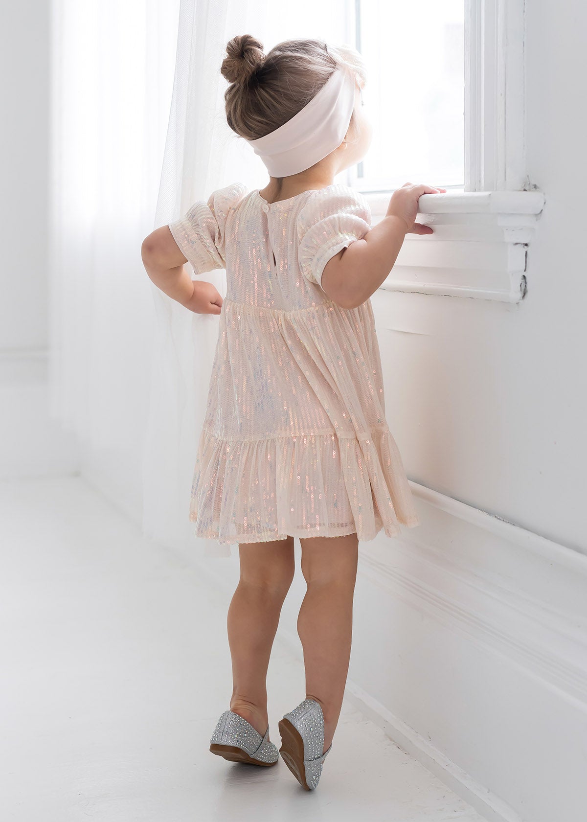A young girl wears the Isobella and Chloe Taylor Sequined Toddler Dress, paired with silver shoes and a white headband, standing on tiptoes by a bright window in a softly lit white room, her hair styled in a bun.