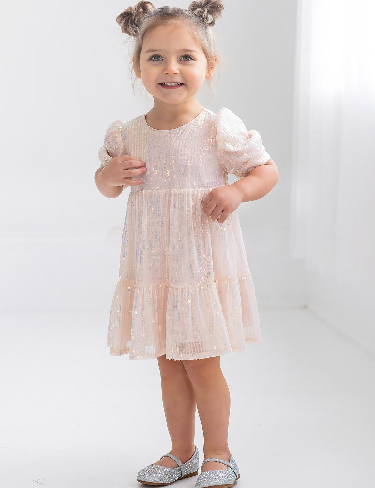 A young girl with light brown hair in two buns smiles while wearing the Isobella and Chloe Taylor Sequined Little Big Girl Dress, featuring puffy sleeves, in a bright white room with silver shoes.