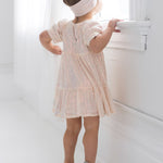 A young girl in the Isobella and Chloe Taylor Sequined Little Big Girl Dress, sparkly silver shoes, and a white headband stands on tiptoe gazing out a bright window in a white room.