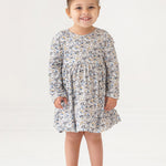 A young girl with brown hair styled in a top bun smiles while standing. She wears the Mabel and Honey Hazelnut Dress, a girls floral long-sleeve dress in light blue, beige, and cream, with beige shoes against a white background.