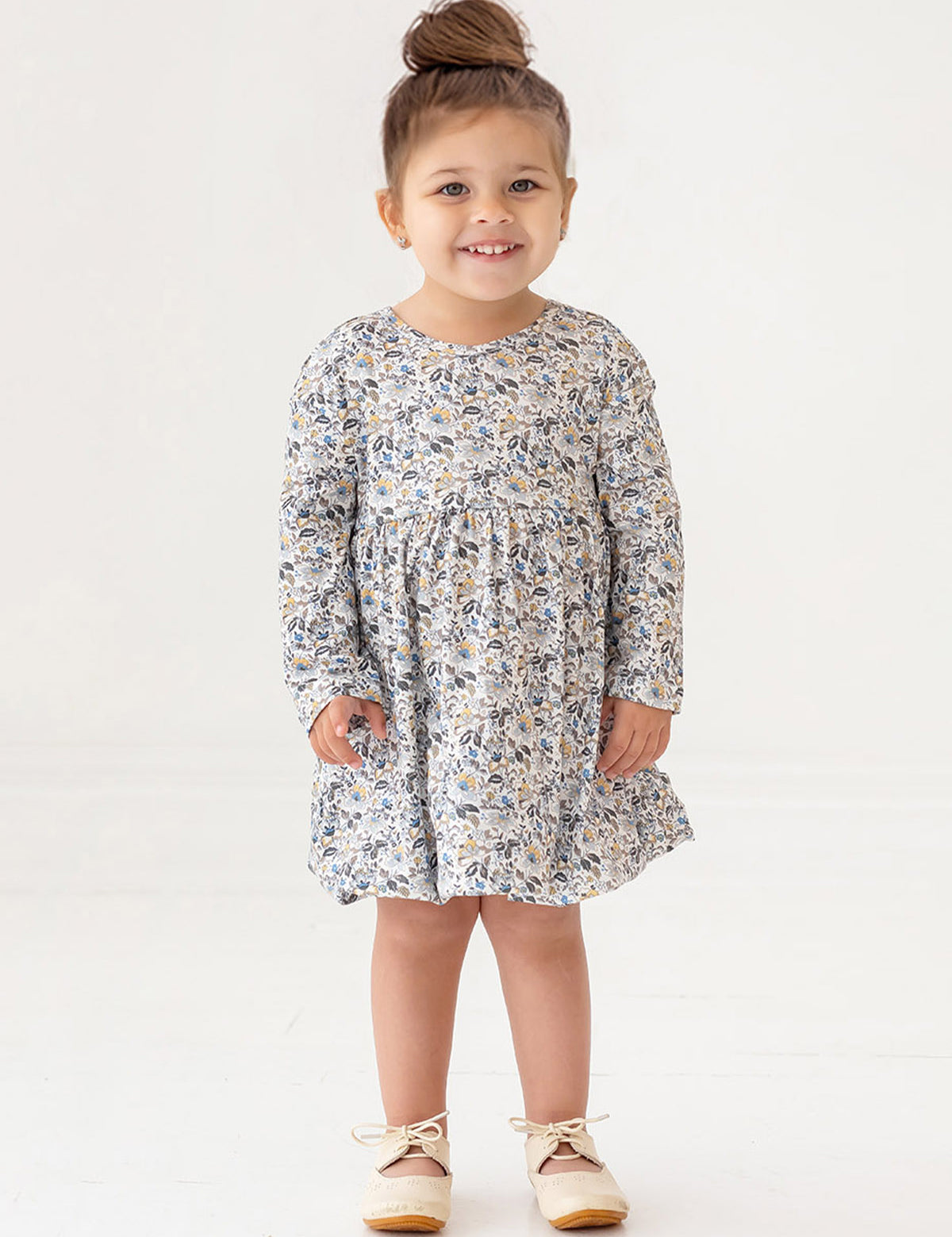 A young girl with brown hair styled in a top bun smiles while standing. She wears the Mabel and Honey Hazelnut Dress, a girls floral long-sleeve dress in light blue, beige, and cream, with beige shoes against a white background.