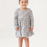 A young girl with brown hair styled in a top bun smiles while standing. She wears the Mabel and Honey Hazelnut Dress, a girls floral long-sleeve dress in light blue, beige, and cream, with beige shoes against a white background.