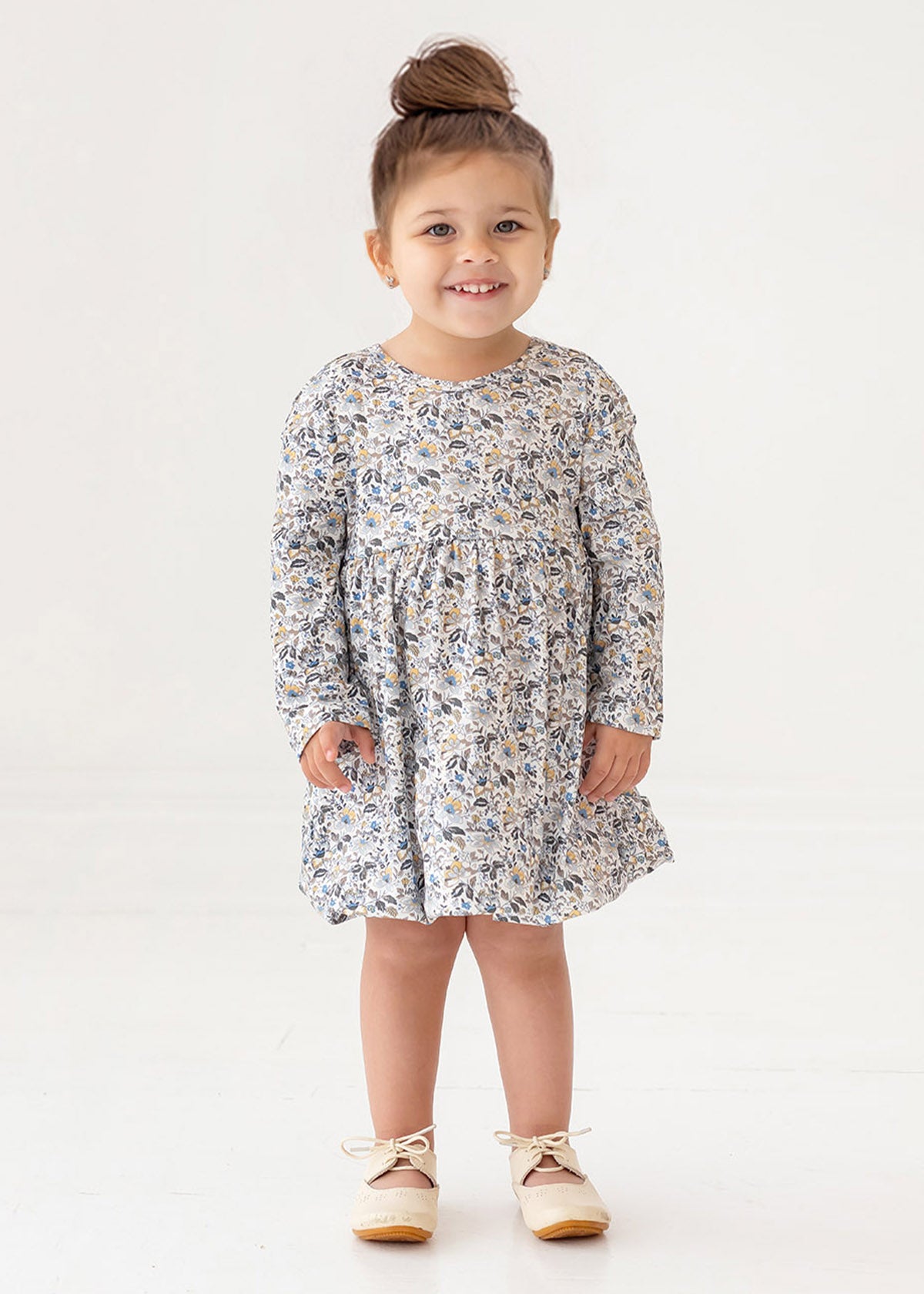 A young girl with brown hair styled in a top bun smiles while standing. She wears the Mabel and Honey Hazelnut Dress, a girls floral long-sleeve dress in light blue, beige, and cream, with beige shoes against a white background.