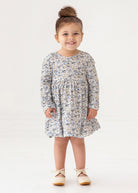 A young girl with brown hair styled in a top bun smiles while standing. She wears the Mabel and Honey Hazelnut Dress, a girls floral long-sleeve dress in light blue, beige, and cream, with beige shoes against a white background.