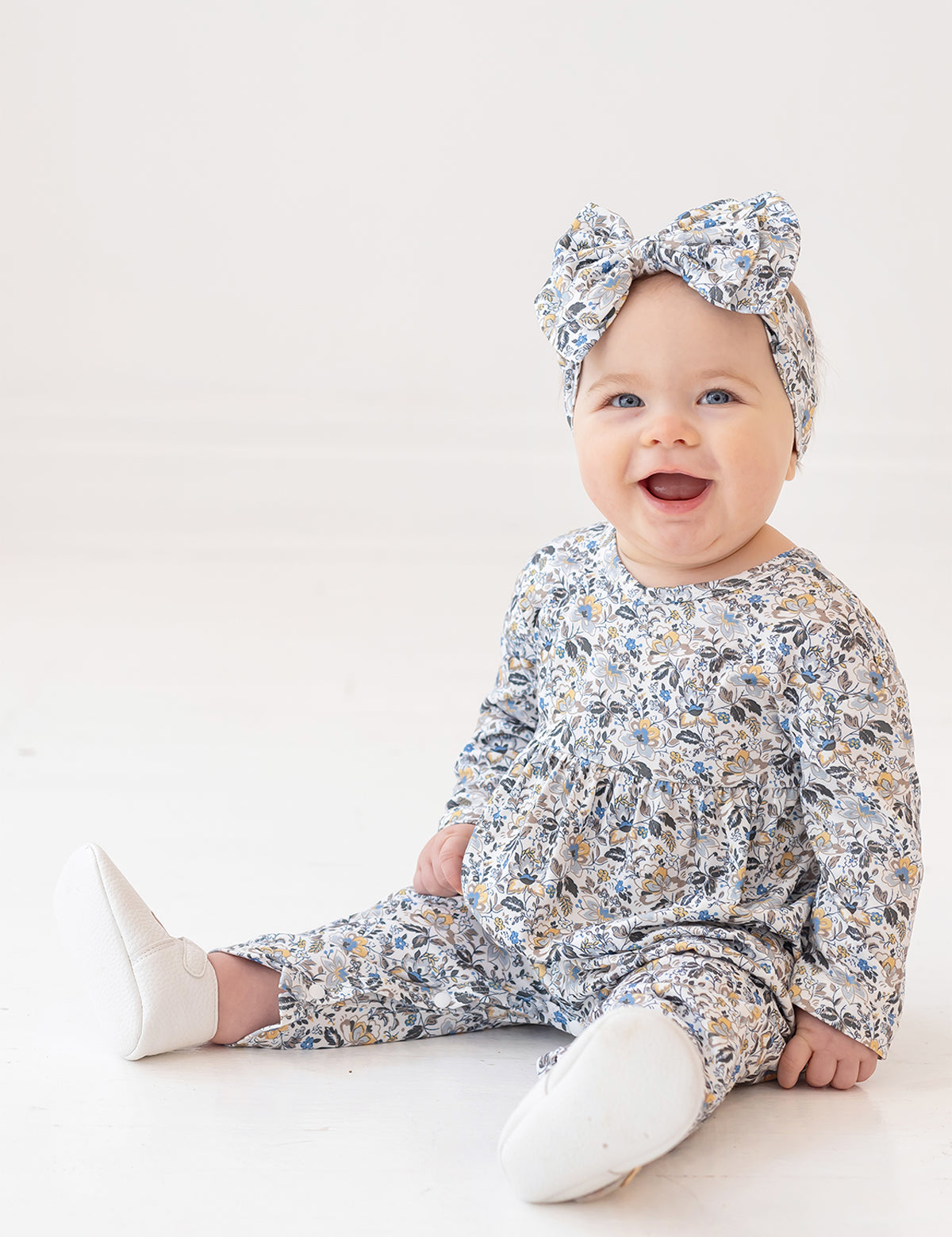 A smiling baby girl wears the Hazelnut Romper by Mabel and Honey with a matching headband, sitting on a white floor against a plain white background.