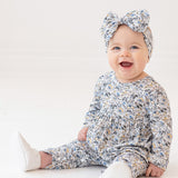 A smiling baby girl wears the Hazelnut Romper by Mabel and Honey with a matching headband, sitting on a white floor against a plain white background.