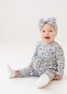 A smiling baby girl wears the Hazelnut Romper by Mabel and Honey with a matching headband, sitting on a white floor against a plain white background.