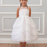 A young girl with braided hair smiles in the Mabel and Honey Sleeveless Organza Tiered Dress with a beaded bodice, white shoes, and stands against a light gray paneled wall on a wooden floor.