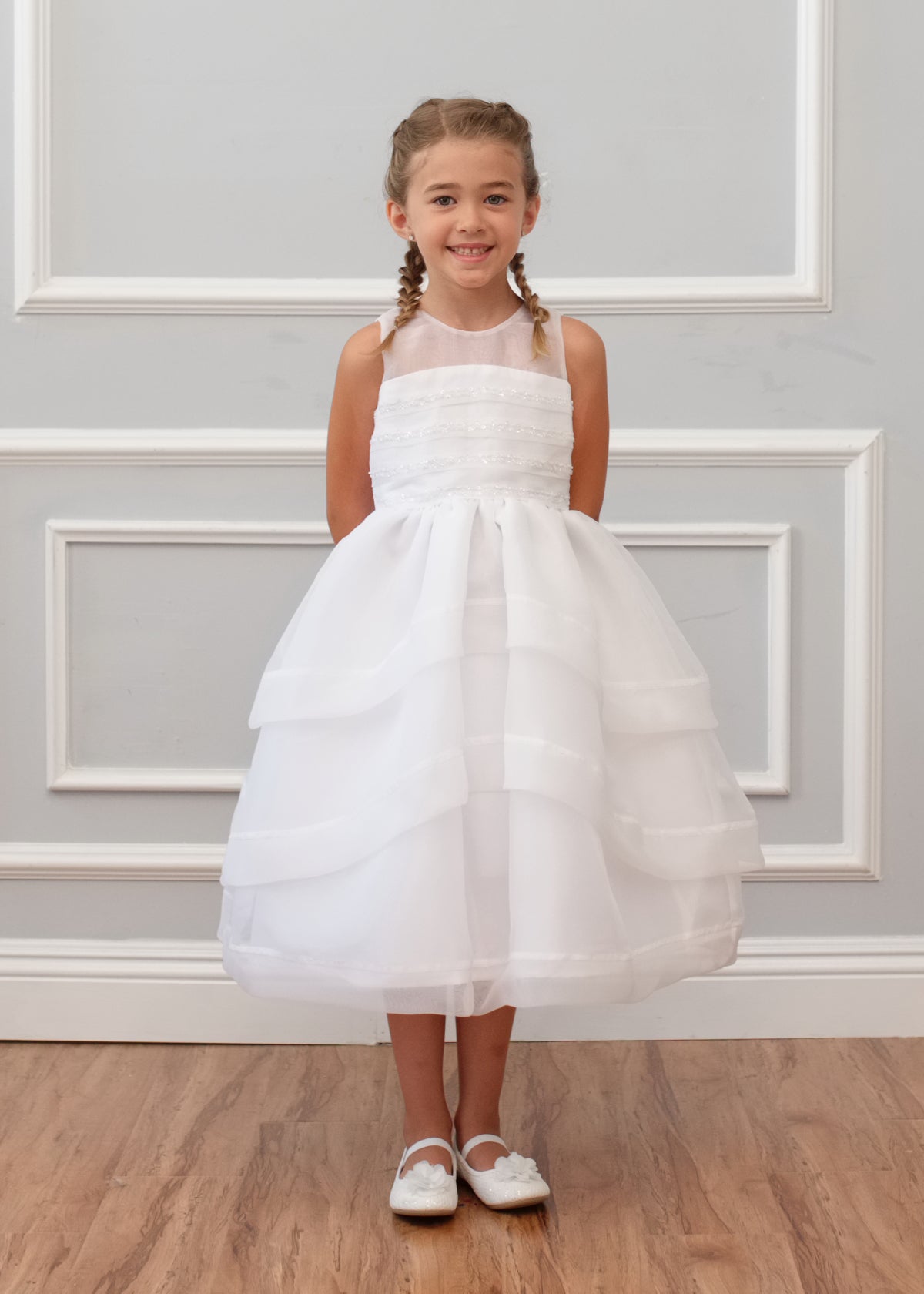 A young girl with braided hair smiles in the Mabel and Honey Sleeveless Organza Tiered Dress with a beaded bodice, white shoes, and stands against a light gray paneled wall on a wooden floor.