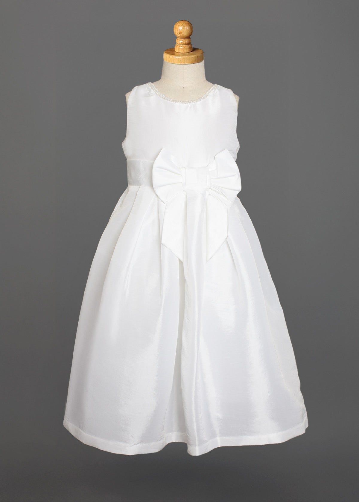 The Taffeta Bow Communion Dress w/ Beaded Pearl Neckline by Mabel and Honey is a sleeveless white girls’ dress with a full skirt, pearl-trimmed neckline, and large waist bow, shown on a dress form against a plain gray background.
