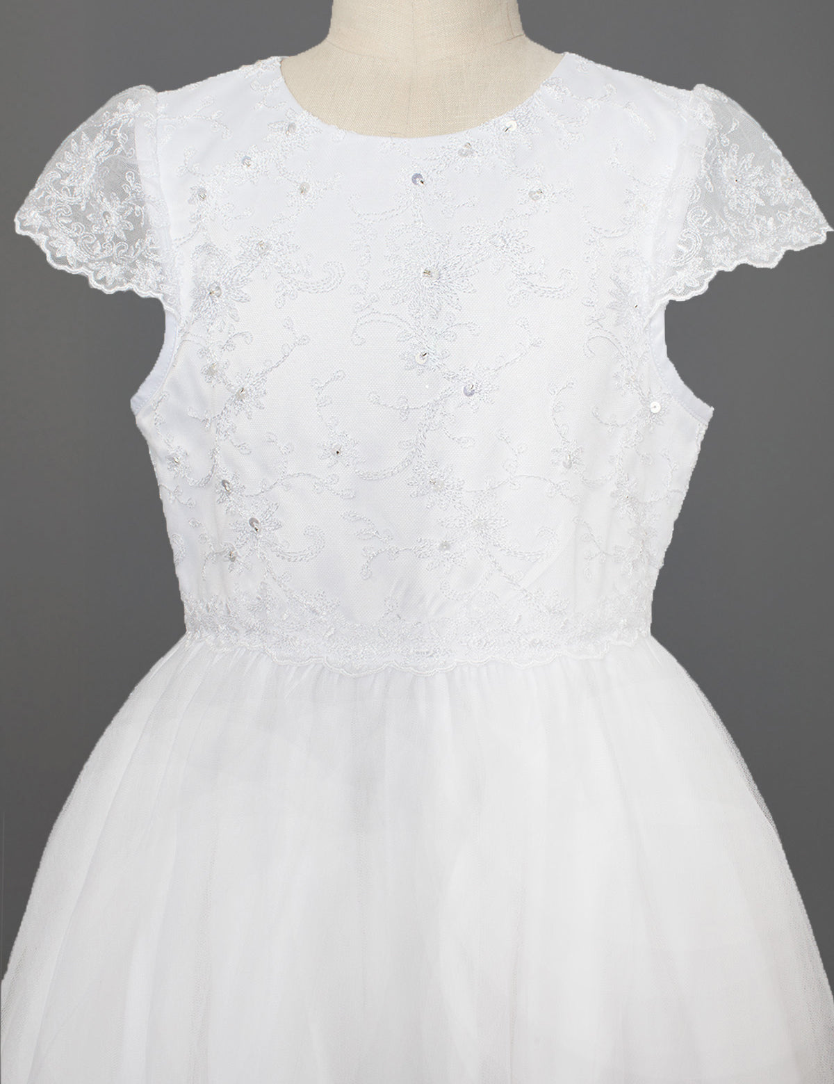 The Mabel + Honey Scalloped Detail on Waist and Sleeve Dress is a white First Communion dress with a lace bodice, delicate embroidery and sequins, cap sleeves, and a full organza tulle skirt. Shown on a mannequin against a gray background.