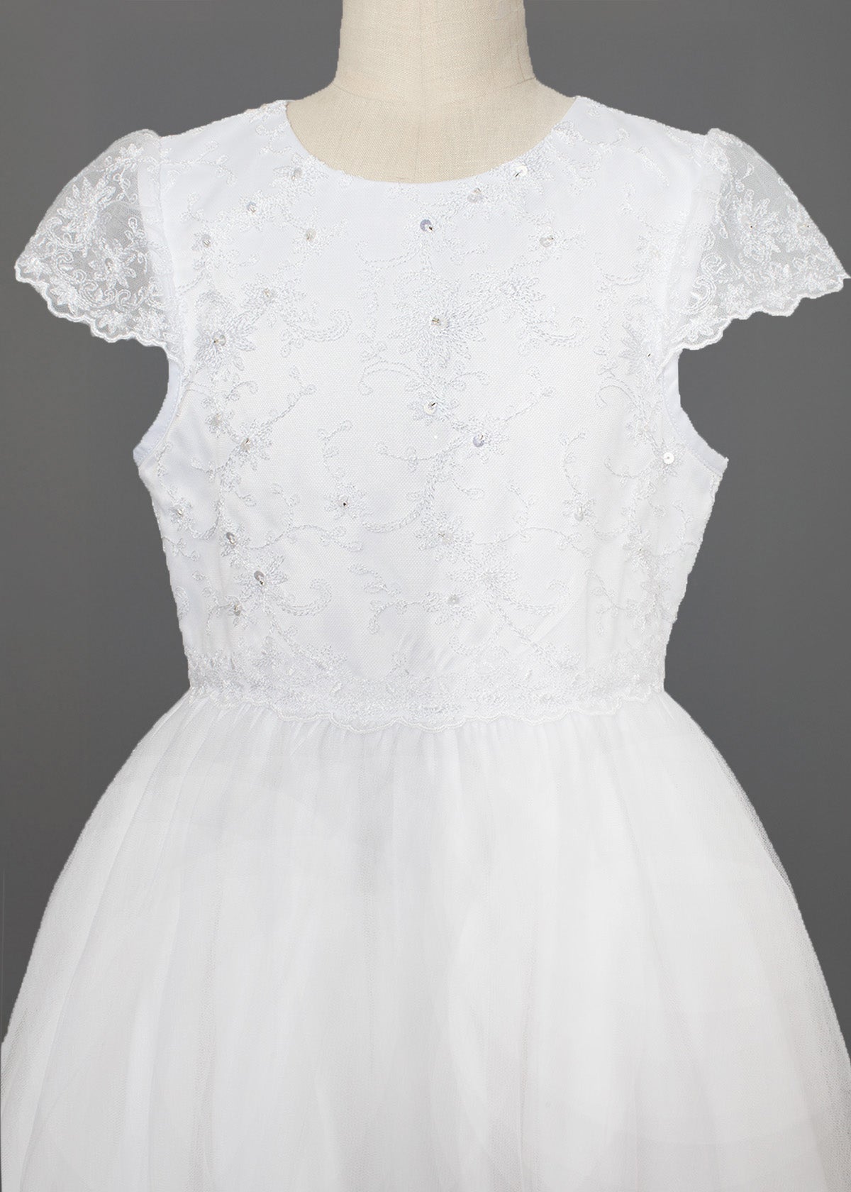 The Mabel + Honey Scalloped Detail on Waist and Sleeve Dress is a white First Communion dress with a lace bodice, delicate embroidery and sequins, cap sleeves, and a full organza tulle skirt. Shown on a mannequin against a gray background.