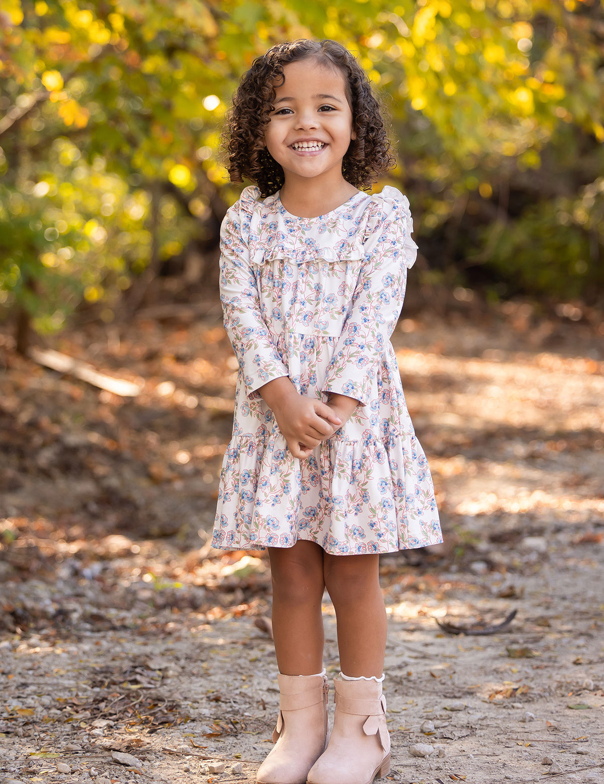 A young girl with curly hair smiles outdoors on a dirt path, wearing the light floral Sadie Dress by Mabel and Honey with pink boots, while sunlight filters through green and yellow trees in the background.