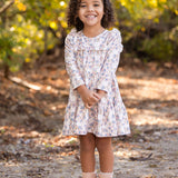 A young girl with curly hair smiles outdoors on a dirt path, wearing the light floral Sadie Dress by Mabel and Honey with pink boots, while sunlight filters through green and yellow trees in the background.