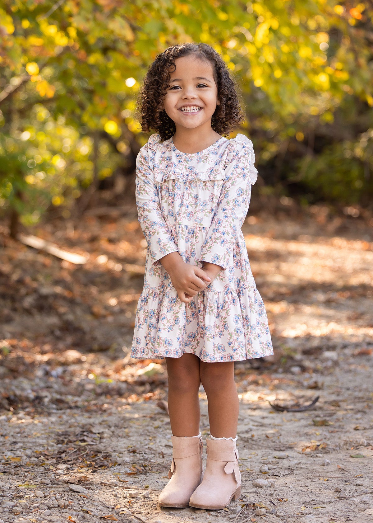 A young girl with curly hair smiles outdoors on a dirt path, wearing the light floral Sadie Dress by Mabel and Honey with pink boots, while sunlight filters through green and yellow trees in the background.