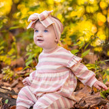 A baby wearing the Mabel and Honey Candy Stripe Romper with ruffled shoulders and a matching headband sits among fallen leaves, surrounded by yellow and green foliage, looking up and smiling.