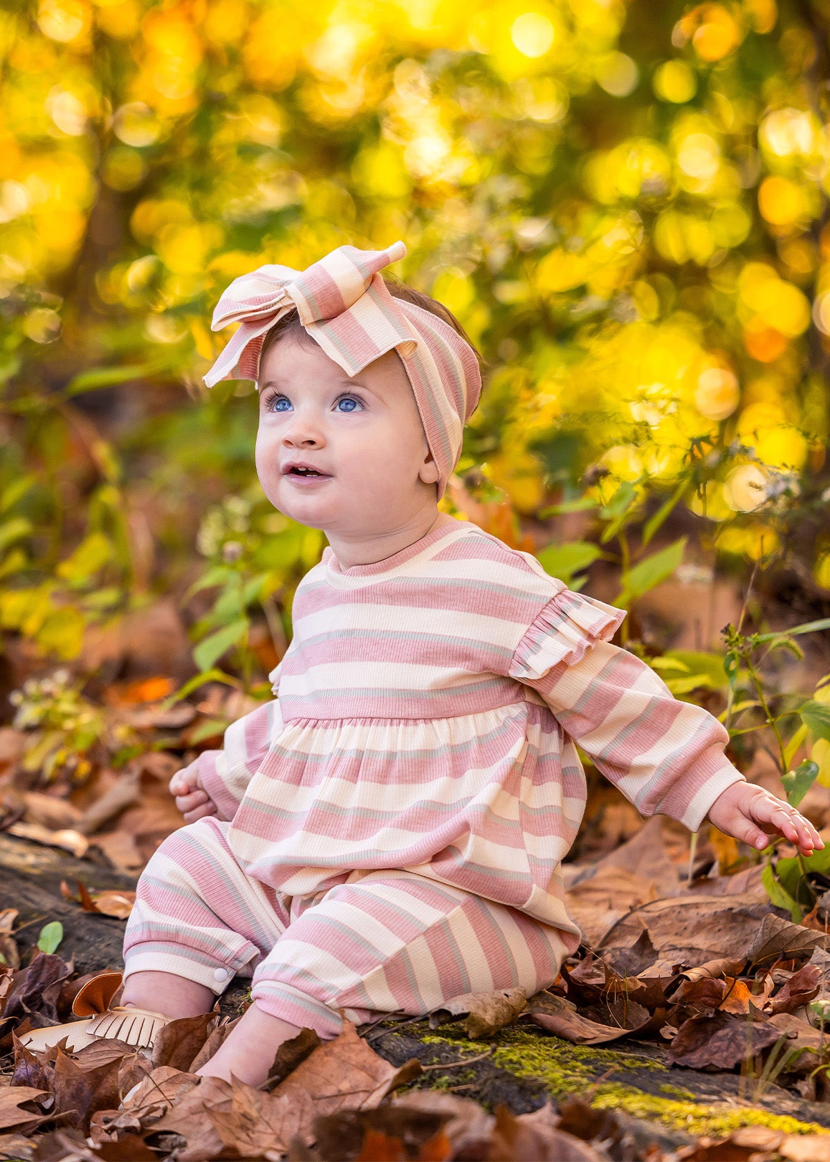 A baby wearing the Mabel and Honey Candy Stripe Romper with ruffled shoulders and a matching headband sits among fallen leaves, surrounded by yellow and green foliage, looking up and smiling.