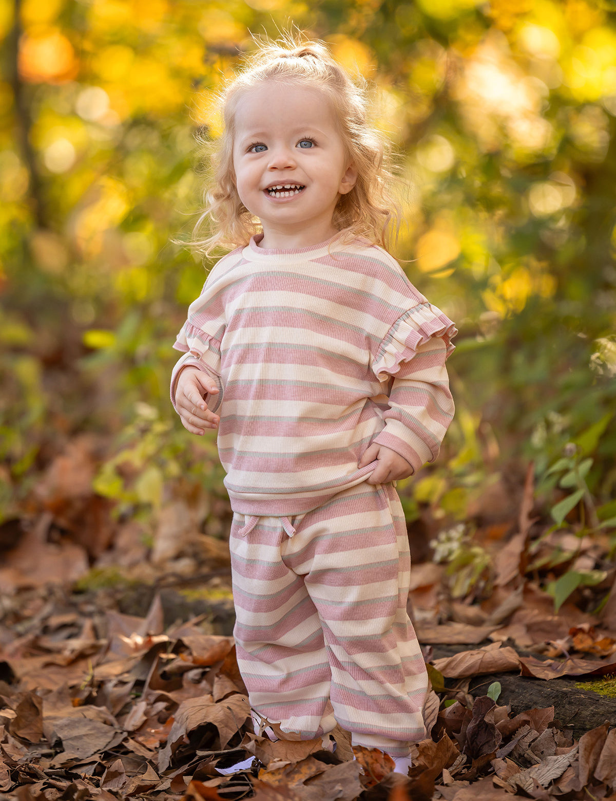 A smiling toddler with curly blonde hair stands among autumn leaves, wearing the cozy Mabel and Honey Candy Stripe Two Piece Set with ruffled sleeves that adds playful charm to her outdoor style.