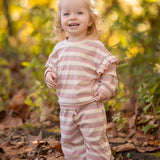 A smiling toddler with curly blonde hair stands among autumn leaves, wearing the cozy Mabel and Honey Candy Stripe Two Piece Set with ruffled sleeves that adds playful charm to her outdoor style.