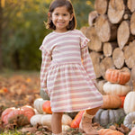 A young girl beams outdoors in the Mabel and Honey Candy Stripe Dress, surrounded by stacked logs and pumpkins with autumn trees behind her—perfectly capturing the essence of fall.