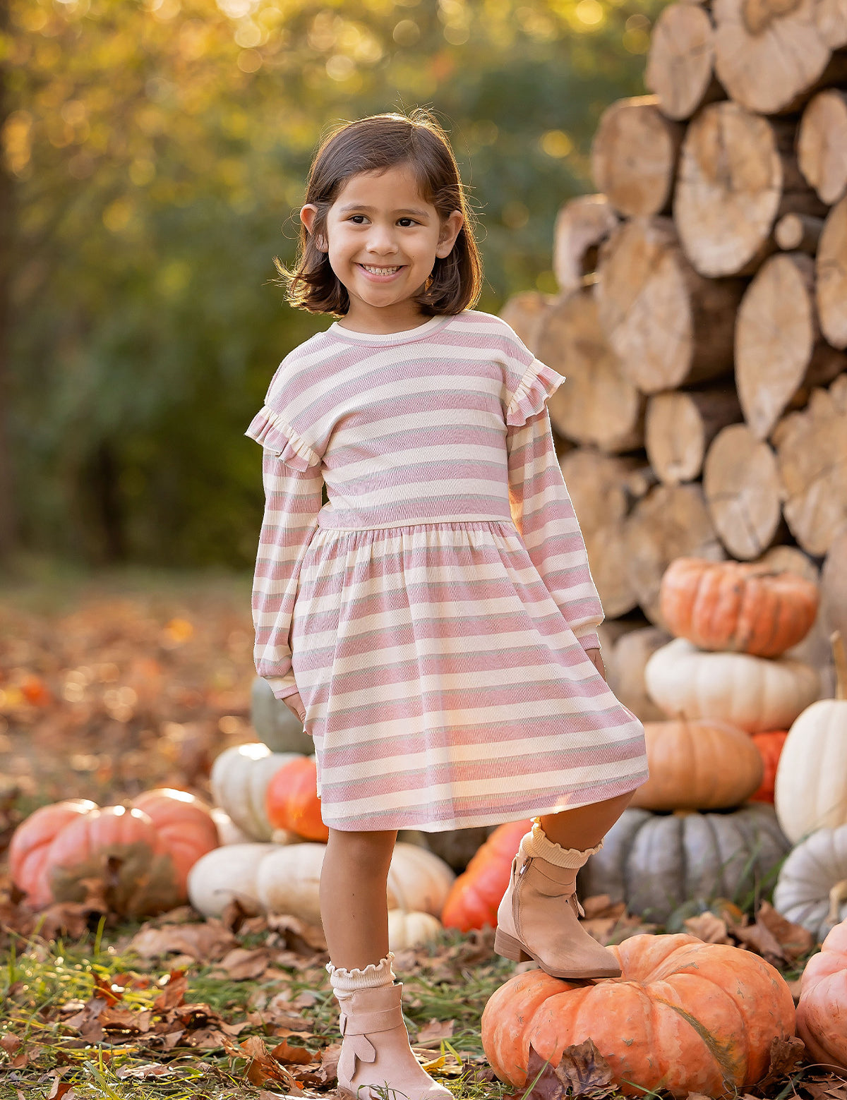 A young girl beams outdoors in the Mabel and Honey Candy Stripe Dress, surrounded by stacked logs and pumpkins with autumn trees behind her—perfectly capturing the essence of fall.