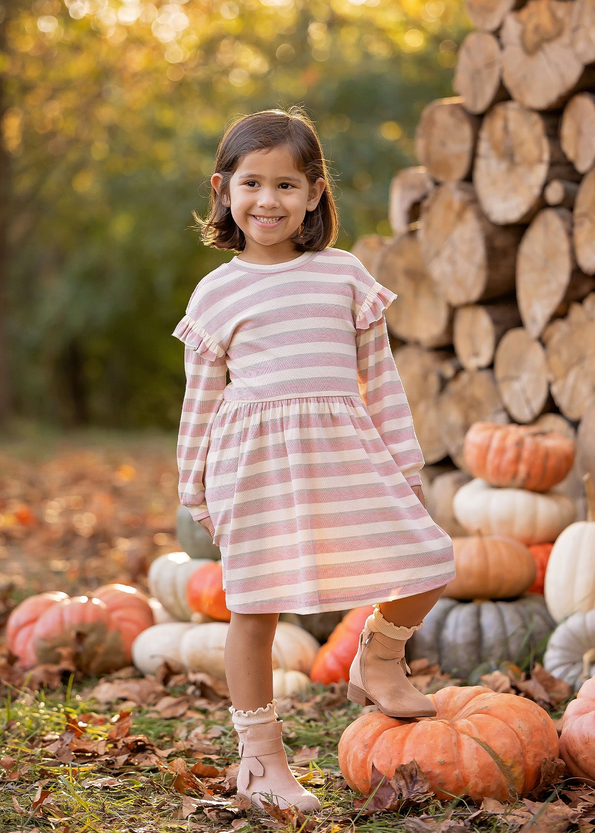A young girl beams outdoors in the Mabel and Honey Candy Stripe Dress, surrounded by stacked logs and pumpkins with autumn trees behind her—perfectly capturing the essence of fall.