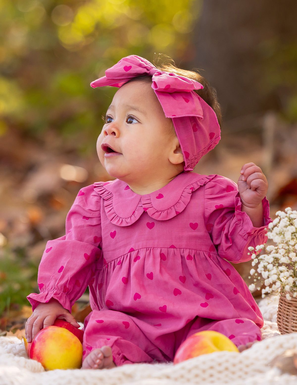 A baby in the Mabel and Honey Perfect in Pink Romper with a matching headband sits outdoors on a blanket, surrounded by apples and flowers, gazing to the side.
