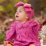 A baby in the Mabel and Honey Perfect in Pink Romper with a matching headband sits outdoors on a blanket, surrounded by apples and flowers, gazing to the side.