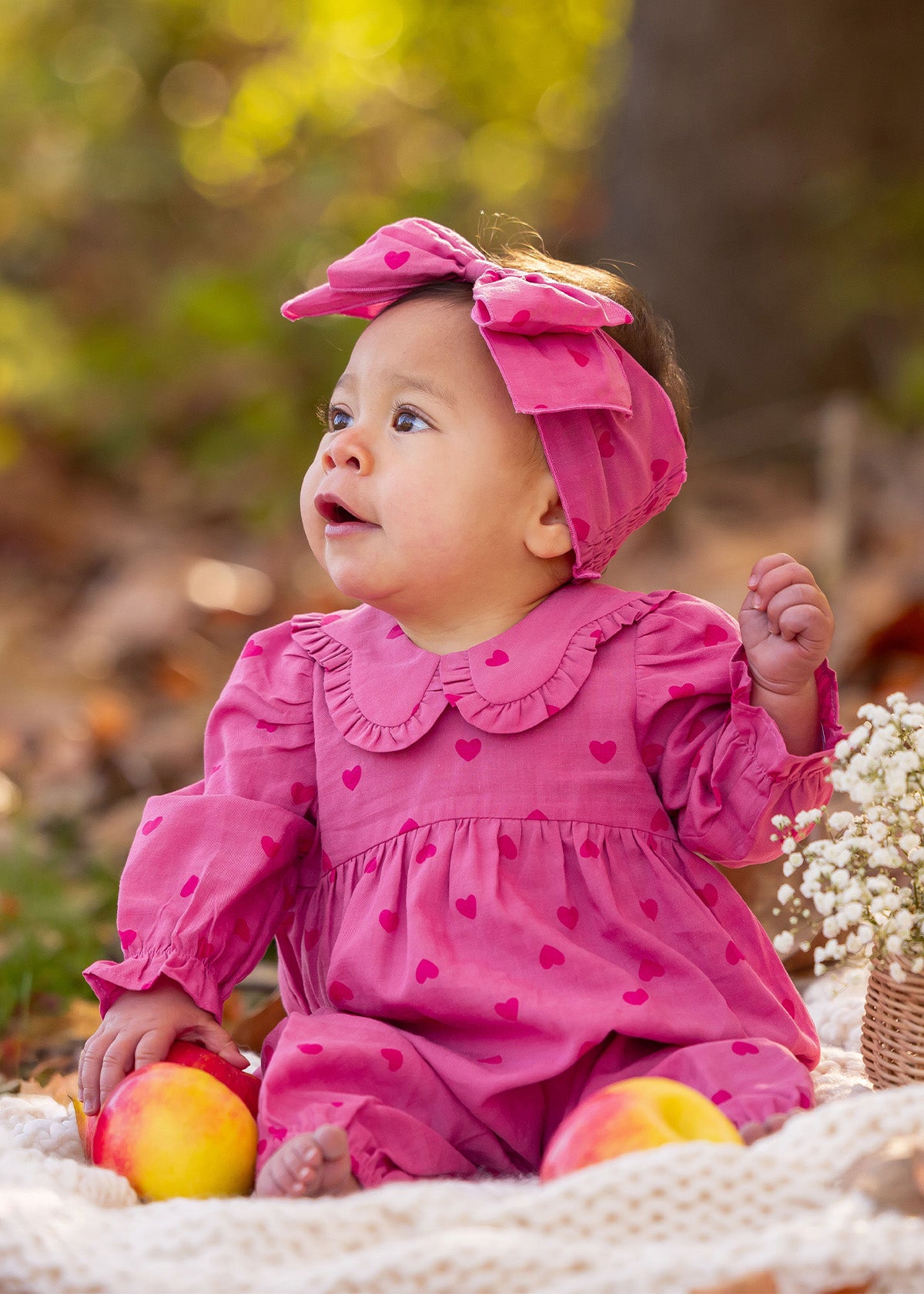 A baby in the Mabel and Honey Perfect in Pink Romper with a matching headband sits outdoors on a blanket, surrounded by apples and flowers, gazing to the side.