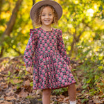 A young girl with curly hair smiles outdoors on fallen leaves, wearing the Mabel and Honey Perfect in Pink Dress with ruffle details, a wide-brim hat, white socks, and tan shoes. Sunlight filters through the trees behind her.