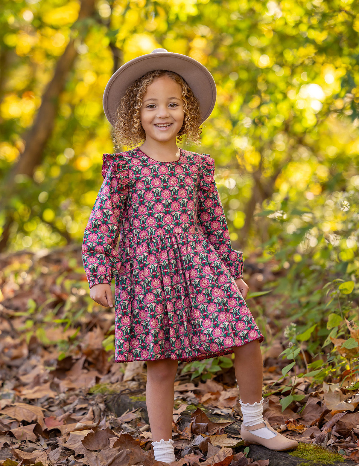 A young girl with curly hair smiles outdoors on fallen leaves, wearing the Mabel and Honey Perfect in Pink Dress with ruffle details, a wide-brim hat, white socks, and tan shoes. Sunlight filters through the trees behind her.