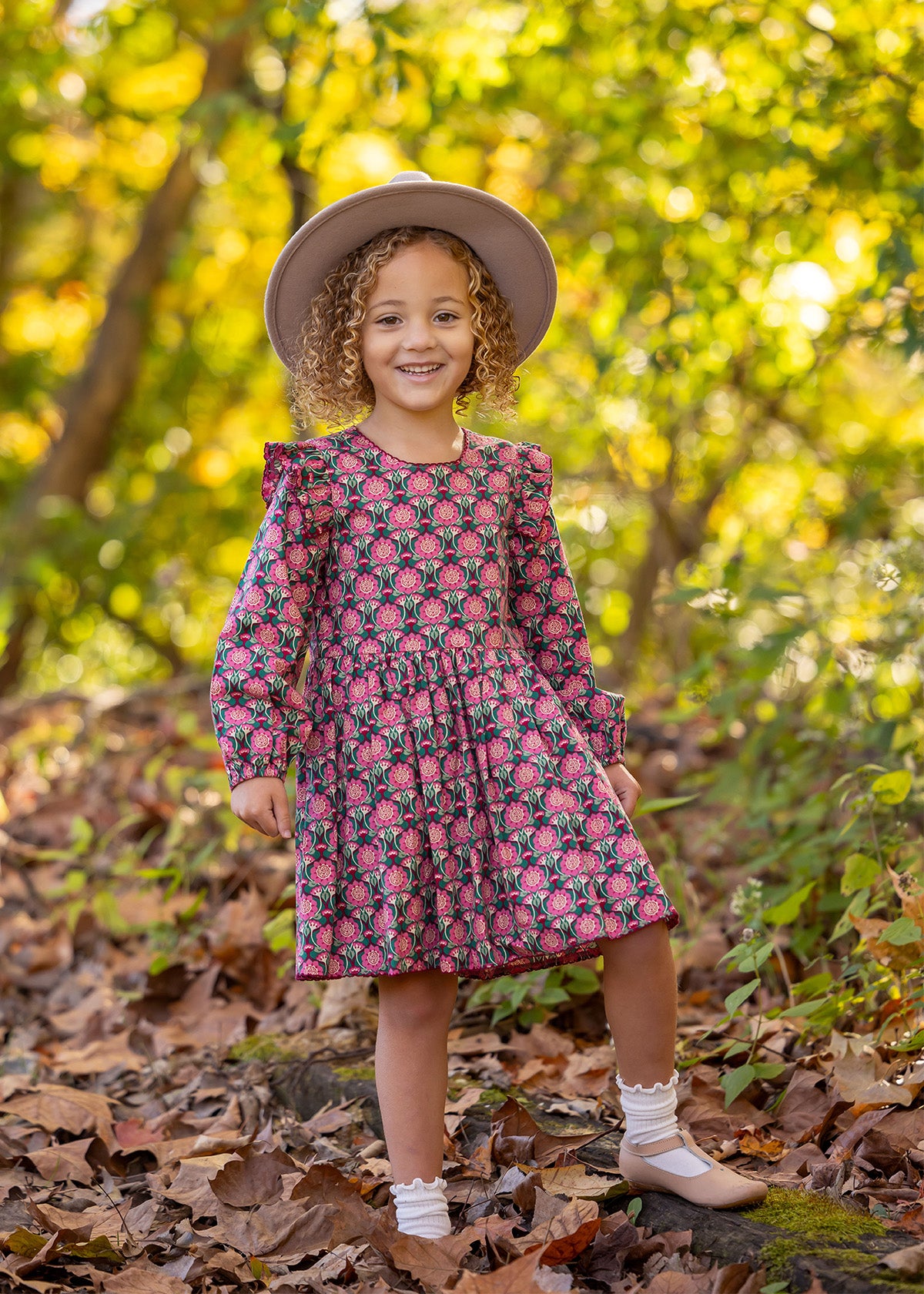 A young girl with curly hair smiles outdoors on fallen leaves, wearing the Mabel and Honey Perfect in Pink Dress with ruffle details, a wide-brim hat, white socks, and tan shoes. Sunlight filters through the trees behind her.