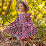 A young child smiles outdoors, wearing the Mabel and Honey Perfect in Pink Dress with ruffle details, white socks, brown shoes, and a light hat, standing among fallen leaves in a sunlit woodland setting.