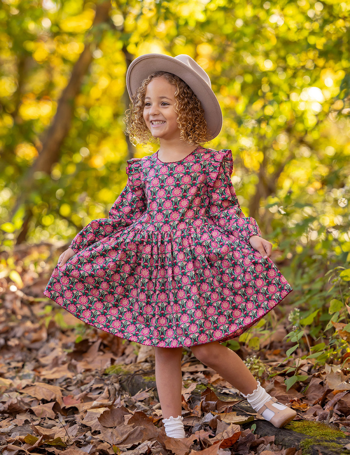 A young child smiles outdoors, wearing the Mabel and Honey Perfect in Pink Dress with ruffle details, white socks, brown shoes, and a light hat, standing among fallen leaves in a sunlit woodland setting.