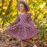 A young child smiles outdoors, wearing the Mabel and Honey Perfect in Pink Dress with ruffle details, white socks, brown shoes, and a light hat, standing among fallen leaves in a sunlit woodland setting.