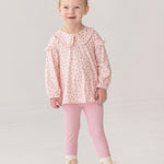 A young child smiles in the Mabel and Honey Primrose Two Piece Set—featuring a ruffled, long-sleeved floral top and pink leggings—paired with cream lace-up boots against a plain light background.