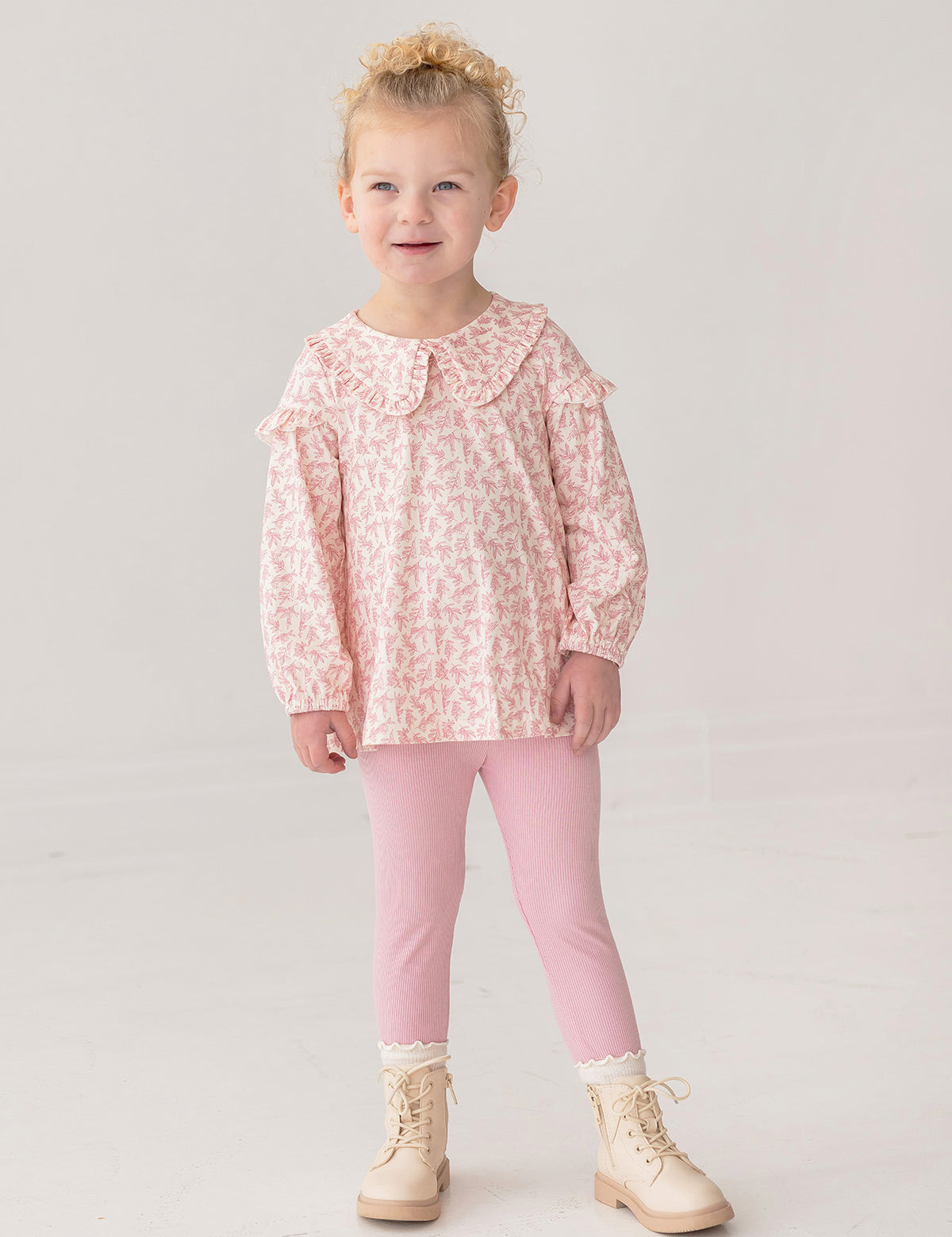 A young child smiles in the Mabel and Honey Primrose Two Piece Set—featuring a ruffled, long-sleeved floral top and pink leggings—paired with cream lace-up boots against a plain light background.