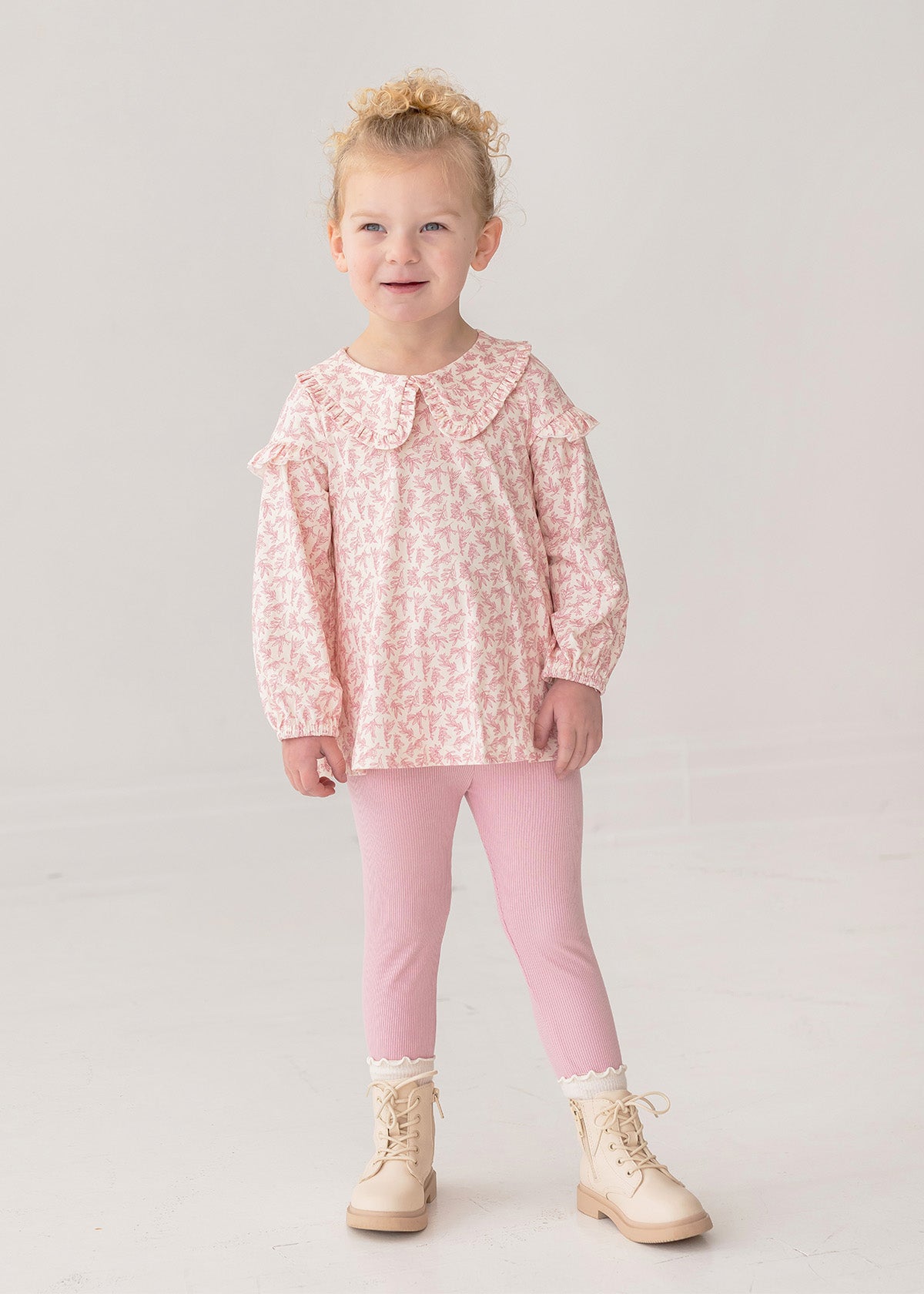 A young child smiles in the Mabel and Honey Primrose Two Piece Set—featuring a ruffled, long-sleeved floral top and pink leggings—paired with cream lace-up boots against a plain light background.