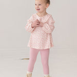 A young girl with curly blonde hair in a bun smiles while wearing the Mabel and Honey Primrose Two Piece Set—featuring a floral top and matching pink leggings—an adorable baby girl outfit, set against a plain white background.