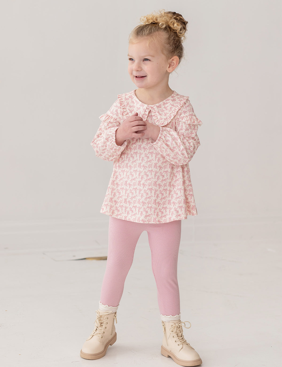 A young girl with curly blonde hair in a bun smiles while wearing the Mabel and Honey Primrose Two Piece Set—featuring a floral top and matching pink leggings—an adorable baby girl outfit, set against a plain white background.