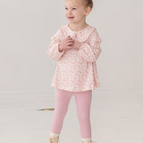 A young girl with curly blonde hair in a bun smiles while wearing the Mabel and Honey Primrose Two Piece Set—featuring a floral top and matching pink leggings—an adorable baby girl outfit, set against a plain white background.